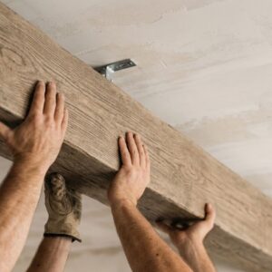 A close-up shot of the hands of two DIYers attempting to lift and align a heavy, textured faux wood decorative beam toward a mounting bracket on a ceiling.