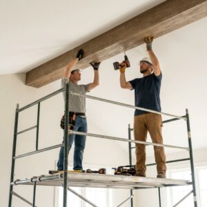 Two professional installers wearing safety gear on a scaffolding securely mounting a faux wood beam to a high ceiling.